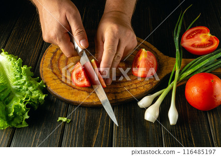 Preparing a salad from fresh vegetables. Cutting a tomato with a knife in the hands of a chef for a vegetarian dish 116485197
