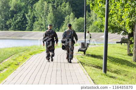 Two male police officers with black uniforms seen from behind walking in the city park  116485870