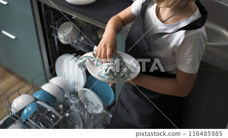 In the kitchen, a young girl wearing an apron wipes dishes that came out of the dishwasher, household duties 116485985