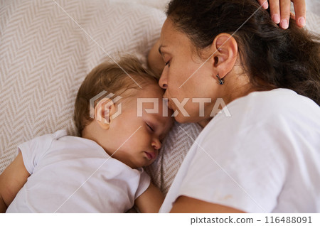 Loving mother kissing her sleeping baby on the forehead while lying together on a cozy bed 116488091