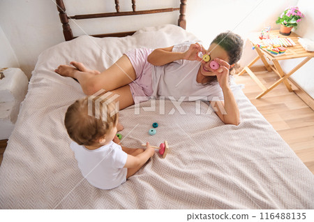 Mother and baby enjoying playful moments on bed with wooden toys in cozy bedroom 116488135