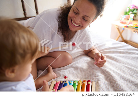 Happy mother playing with child on bed with colorful xylophone, enjoying family time Happy mother playing with child on bed with colorful xylophone, enjoying family time 116488164