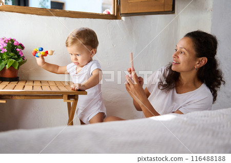 Mother photographing her baby playing with a colorful toy near a window 116488188