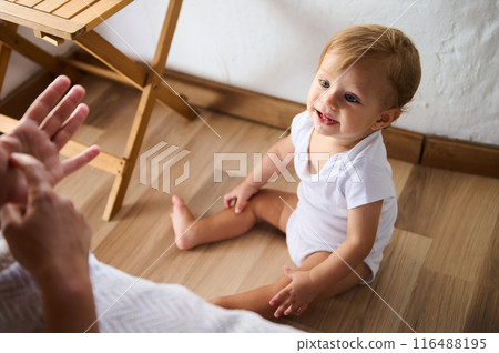 Adorable baby in white onesie sitting on the wooden floor and smiling while interacting with a parent 116488195