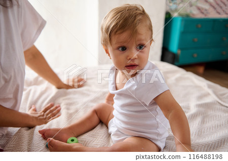 Adorable baby sitting on bed in bright, cozy room, expressing curiosity and wonder Adorable baby sitting on bed in bright, cozy room, expressing curiosity and wonder 116488198
