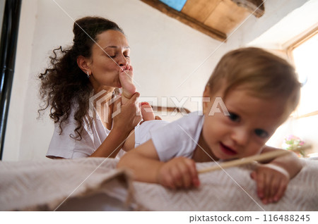 Mother kissing baby's foot while child crawls on bed in cozy home interior Mother kissing baby's foot while child crawls on bed in cozy home interior 116488245