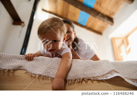 Happy toddler playing with mother on wooden bed in cozy home environment Happy toddler playing with mother on wooden bed in cozy home environment 116488246