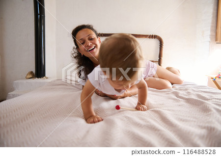 Happy mother and baby playing on bed in cozy room with natural light Happy mother and baby playing on bed in cozy room with natural light 116488528
