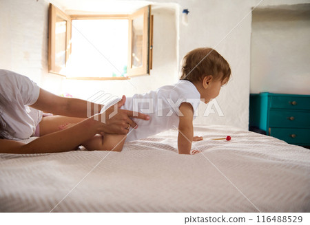 Parent supporting baby learning to crawl on bed in bright bedroom Parent supporting baby learning to crawl on bed in bright bedroom 116488529