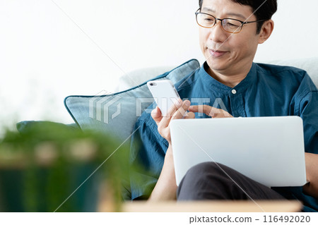 A middle-aged man operating a computer and smartphone on the sofa 116492020