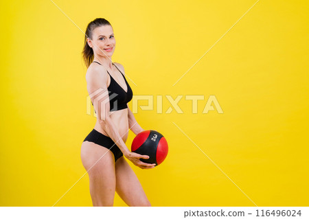 Sportive caucasian cross fit woman with fitness ball, look at camera. Studio dark yellow background 116496024