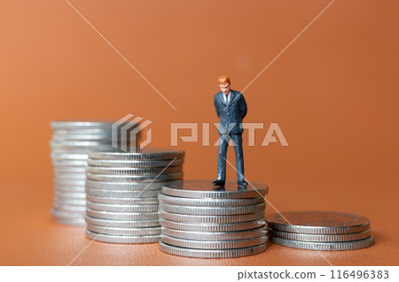 Miniature people A businessman in a suit stands with several stacks of silver coins 116496383