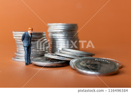 Miniature people A businessman in a suit stands with several stacks of silver coins 116496384