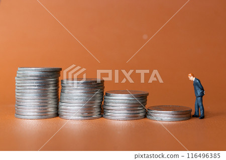 Miniature people A businessman in a suit stands with several stacks of silver coins 116496385