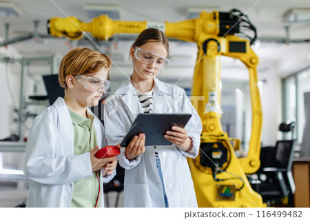 Two classmates standing in robotic laboratory, wearing lab coats and safety eyeglasses. After-school robotics club. Children learning robotics in Elementary school. 116499482