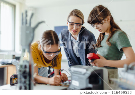 Female teacher helping to girls working on small robot, working together on circuit board in after-school robotics club. Children learning robotics in Elementary school. Girls in science. Female teacher helping to girls working on small robot, working together on circuit board in after-school robotics club. Children learning robotics in Elementary school. Girls in science. 116499533