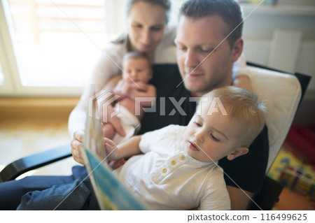 Nuclear family with neborn baby daughter reading book with older son. Mom and dad spending time with kids in room. Nuclear family with neborn baby daughter reading book with older son. Mom and dad spending time with kids in room. 116499625