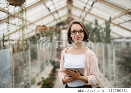 Small greenhouse business. Businesswoman selling flowers and seedlings, standing in greenhouse, looking at camera, smiling. Small greenhouse business. Businesswoman selling flowers and seedlings, standing in greenhouse, looking at camera, smiling. 116500281