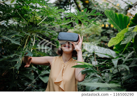 Handsome businessman standing in botanical garden, in the middle of lush green foliage, using VR headset. Handsome businessman standing in botanical garden, in the middle of lush green foliage, using VR headset. 116500290