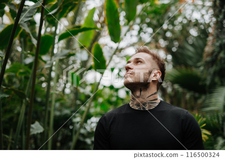 Handsome man with tattoo standing in botanical garden, in the middle of lush green foliage, looking at camera. Handsome man with tattoo standing in botanical garden, in the middle of lush green foliage, looking at camera. 116500324
