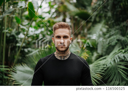 Handsome man with tattoo standing in botanical garden, in the middle of lush green foliage, looking at camera. Handsome man with tattoo standing in botanical garden, in the middle of lush green foliage, looking at camera. 116500325