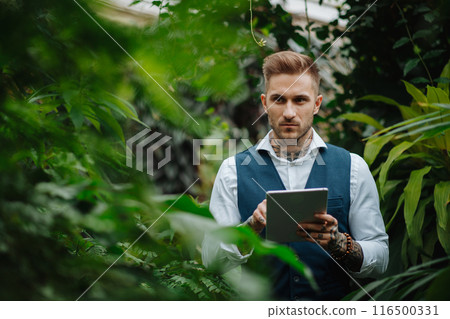 Handsome businessman with tablet, standing in botanical garden, in the middle of lush green foliage, looking at camera. Concept of business corporate social responsibility and ESG. Handsome businessman with tablet, standing in botanical garden, in the middle of lush green foliage, looking at camera. Concept of business corporate social responsibility and ESG. 116500331