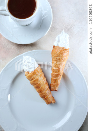 Puff pastry tubes with meringue and a cup of coffee on the table. Sweet Italian dessert on a white plate. Top view. 116500868