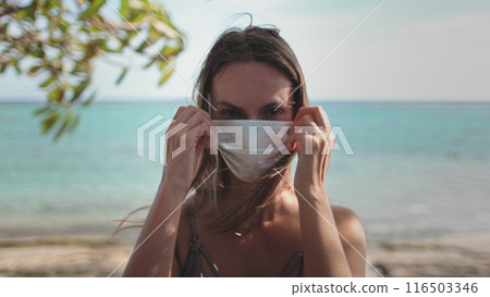 Woman put mask on face close up standing on tropical beach. Long hair caucasian girl on exotic island outdoor lifestyle travel summer holiday vacation. Covid pandemic prevention. Zoom out Woman put mask on face close up standing on tropical beach. Long hair caucasian girl on exotic island outdoor lifestyle travel summer holiday vacation. Covid pandemic prevention. Zoom out 116503346