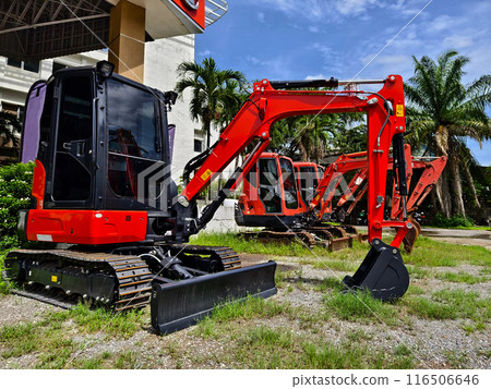 Red large excavators are stationed in a green field in front of a building Red large excavators are stationed in a green field in front of a building 116506646
