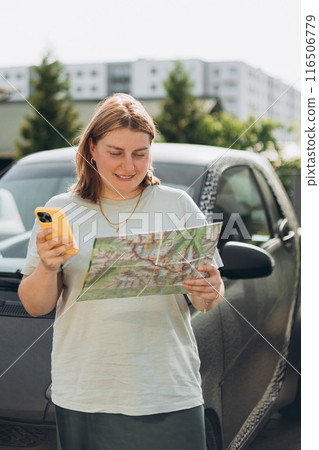 Smiling 30s woman with paper map standing near her automobile on a parking and using a phone outdoors. Urban lifestyle, technology concept. 116506779