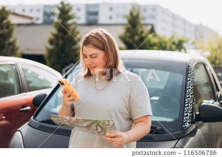 Smiling 30s woman with paper map standing near her automobile on a parking and using a phone outdoors. Urban lifestyle, technology concept. Smiling 30s woman with paper map standing near her automobile on a parking and using a phone outdoors. Urban lifestyle, technology concept. 116506786
