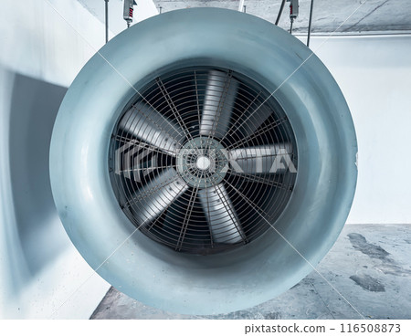 A large, gray, industrial fan is suspended from the ceiling of a concrete building. 116508873