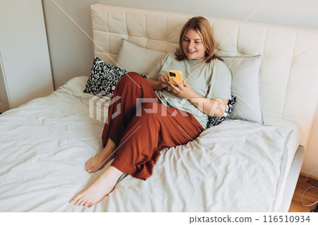 Young woman holding smartphone, using cellphone modern technology, checking cell phone apps, texting, watching video, browsing internet for shopping. Girl sitting on the bed at home. Full length body 116510934