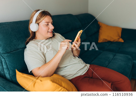 Young 30s woman wear t-shirt and headphones listen music, use mobile cell phone, sits on sofa, stay at home or flat. Concepts of education, work, rest and comfort. 116510936