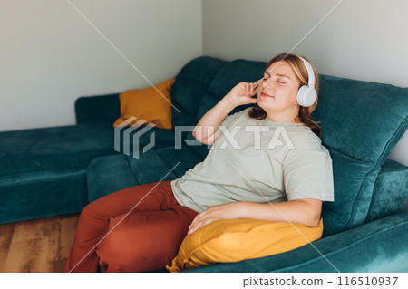Young 30s woman wear t-shirt and headphones listen music, sits on sofa, stay at home or flat. Concepts of education, relax and comfort. Happy woman wearing wireless headphones close eyes 116510937