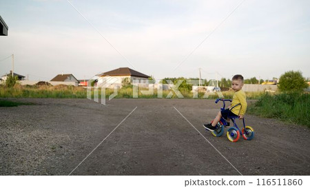 Summer, child, bike on the road. A little boy rides a tricycle. A child in a T-shirt rides a tricycle along the road in a new village. 116511860