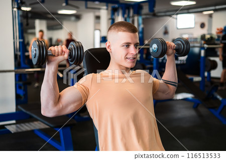 Young bodybuilder lifts dumbbells while sitting on a bench in a gym 116513533