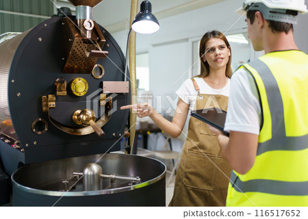 Maintenance engineer or repairman inspecting or checking an automatic coffee roasting machine for maintenance. Repairman talking to female customer about fixing and repairing coffee roasting machine. 116517652