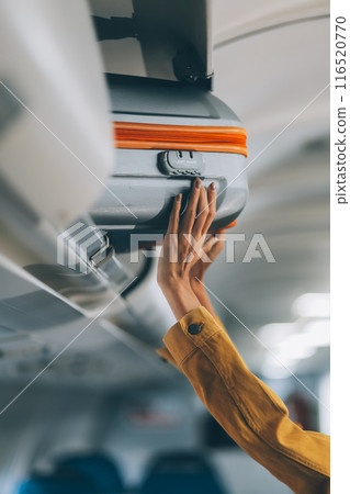 Young Female Tourist Storing Luggage in Overhead Compartment on Airplane During Travel Young Female Tourist Storing Luggage in Overhead Compartment on Airplane During Travel 116520770