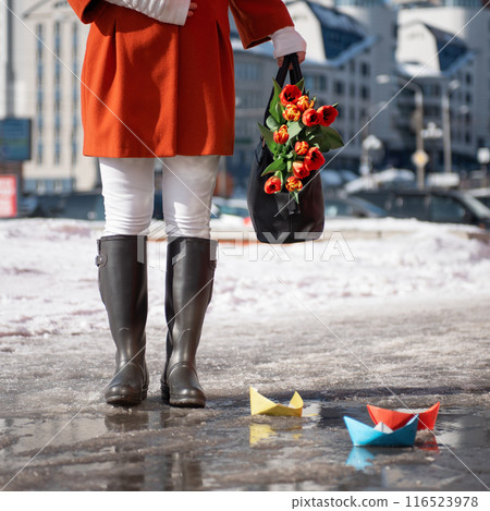 Closeup of woman in a orange coat and rubber boots walking down the street with a bouquet of tulips 116523978