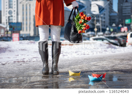 A beautiful young woman in a orange coat and rubber boots walking down the street with a bouquet of tulips A beautiful young woman in a orange coat and rubber boots walking down the street with a bouquet of tulips 116524043