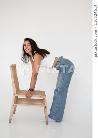 Happy beautiful girl posing with a chair in a new hot look. Wearing blue jeans and white waistcoat Happy beautiful girl posing with a chair in a new hot look. Wearing blue jeans and white waistcoat 116524614