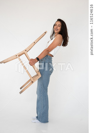 Happy beautiful girl posing with a chair in a new hot look. Wearing blue jeans and white waistcoat 116524616
