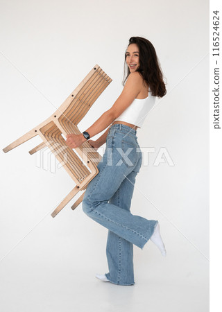Happy beautiful girl posing with a chair in a new hot look. Wearing blue jeans and white waistcoat 116524624