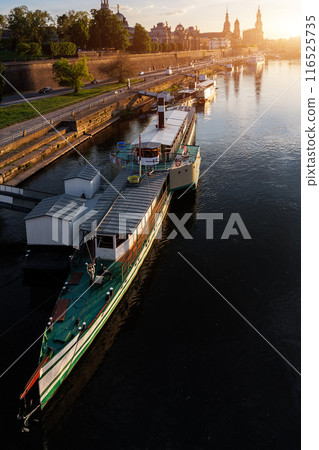 Scenic warm sunset beautiful Dresden city skyline at Elbe River with many steam tourist ship vessel moored at bank. Saxony Germany capital cityscape dusk panoramic view with old Europe architecture Scenic warm sunset beautiful Dresden city skyline at Elbe River with many steam tourist ship vessel moored at bank. Saxony Germany capital cityscape dusk panoramic view with old Europe architecture 116525735