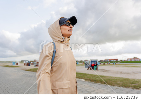 Smiling happy woman walking by Nord sea coast wear beige coat  sunglasses enjoys windy day cloudy sky background. Female person have fun hiking adventure travel at Baltic sea seaside 116525739