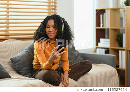 Relaxed young woman with curly hair wearing headphones listening to music, using mobile phone on sofa Relaxed young woman with curly hair wearing headphones listening to music, using mobile phone on sofa 116527634