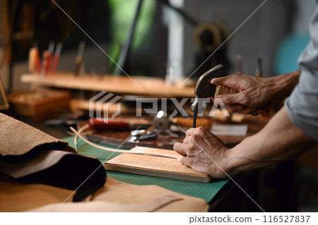 Close up shot of skilled leather craftsman punching a leather detail at workshop small studio Close up shot of skilled leather craftsman punching a leather detail at workshop small studio 116527837