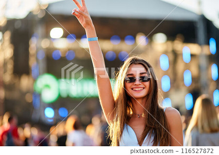 Smiling young woman enjoys summer music festival, raises peace sign, wears sunglasses, wristband, against stage lights. Happy crowd, outdoor concert vibes, live performance event at beach. Smiling young woman enjoys summer music festival, raises peace sign, wears sunglasses, wristband, against stage lights. Happy crowd, outdoor concert vibes, live performance event at beach. 116527872