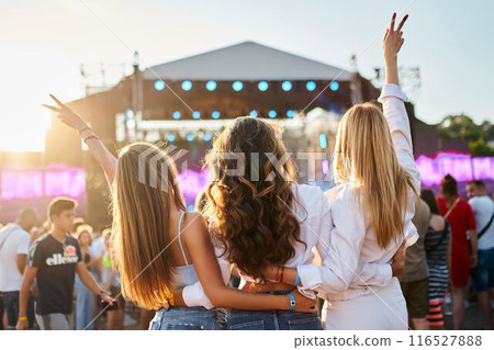 Group of girls at beach music fest, arms raised, enjoying sunset. Friends hug, celebrate, dance in crowd, summer party vibe. Sunlight flares on stage, festival-goers gather, casual outdoor fun. 116527888